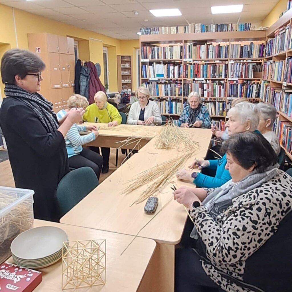 Kūrybinis užsiėmimas Ginkūnų bibliotekoje – dalyvės prie stalo pina dirbinius iš šiaudų.