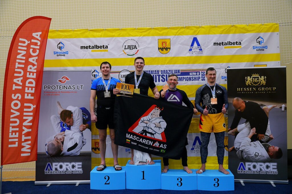 Ankstesnių braziliškojo Jiu-Jitsu varžybų Šiauliuose apdovanojimų akimirka – ant nugalėtojų pakylos su medaliais stovi keturi sportininkai, vienas jų laiko „Mascarenhas Team“ vėliavą.