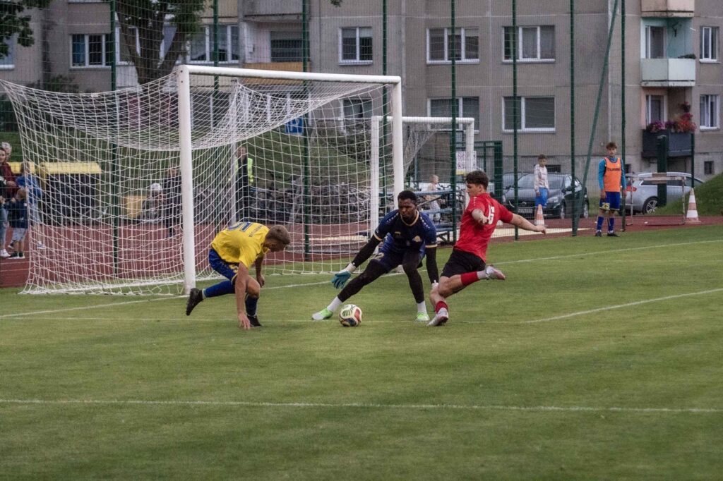 Kuršėnų „Ventos“ futbolininkas atakuoja Plungės „Babrungo B“ vartus, mėgindamas įveikti varžovų gynėją ir vartininką.