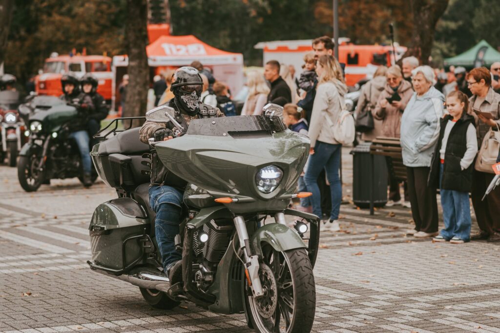 Baikeris motociklu važiuoja per Kuršėnų Jomarko šventę, o pakelėje susirinkę žmonės stebi koloną.