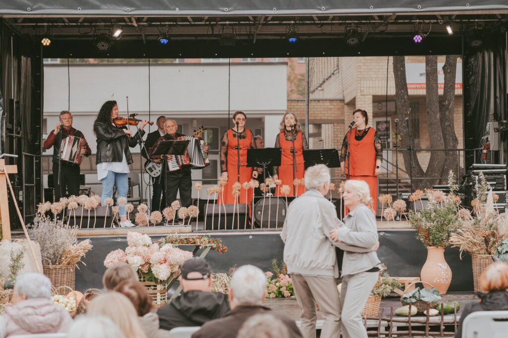 uršėnų Jomarko scenoje koncertuoja muzikantai ir dainininkės, o priešais sceną šoka pora. Scena papuošta rudens gėlėmis ir derliaus gėrybėmis.
