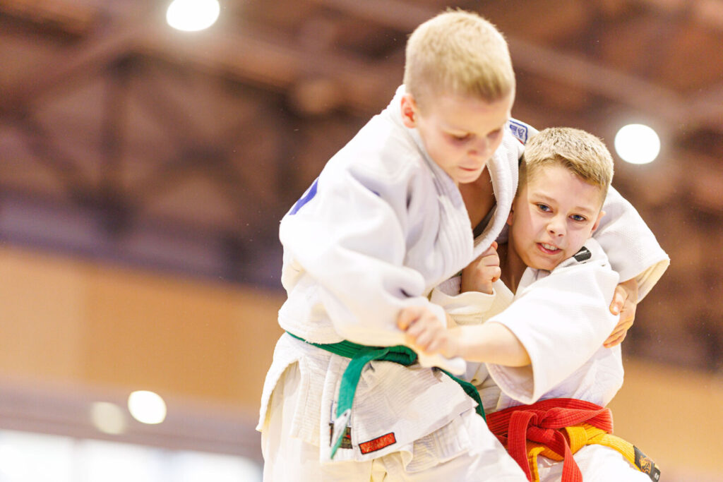 Du jaunieji dziudo sportininkai treniruotės metu atlieka imtynių veiksmą sporto salėje.