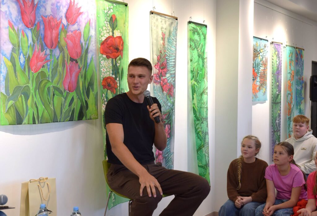 Keliautojas ir turinio kūrėjas Lukas Vainavičius su mikrofonu rankoje pasakoja apie keliones ir patirtus nuotykius Vytauto Vitkausko viešojoje bibliotekoje Kuršėnuose, susirinkusiems vaikams ir kitiems renginio dalyviams.