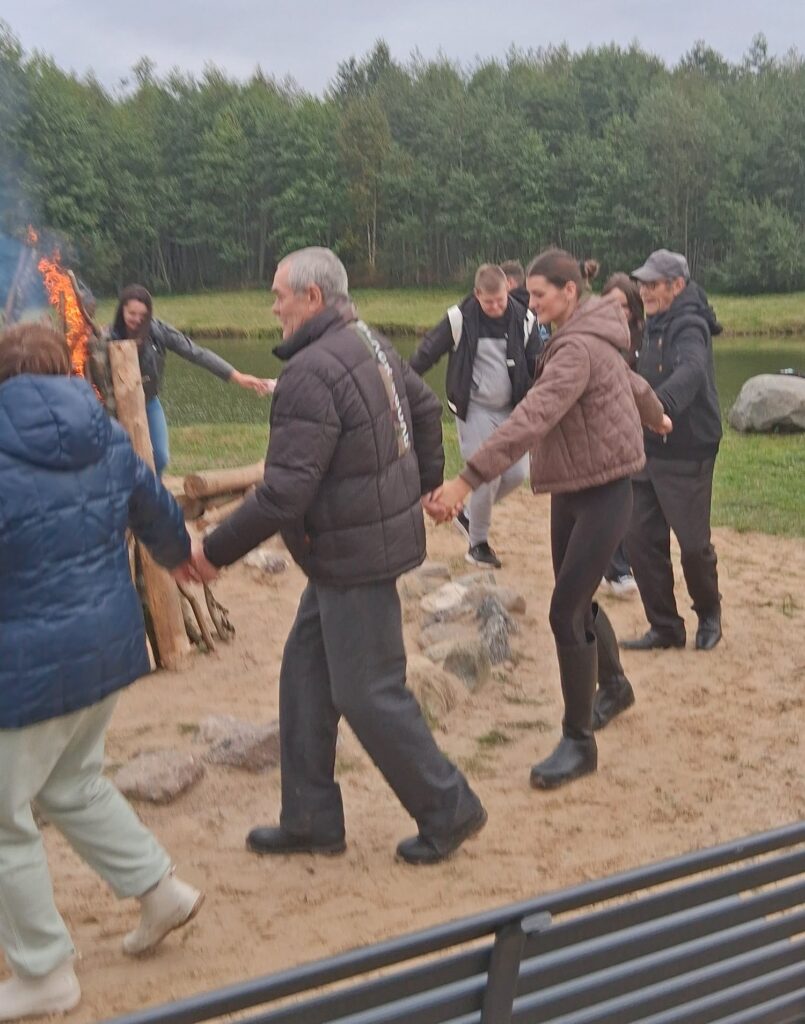 Noreikių bendruomenės šventės dalyviai susikibę rankomis šoka rateliu prie laužo ir tvenkinio.