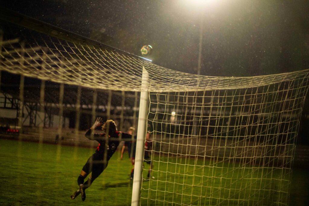 FK „Venta“ ir Vilniaus „Vyčio“ futbolo rungtynių akimirka – vakare, lyjant lietui, kamuolys skrieja virš vartų skersinio, o vartininkas bando jį pasiekti.
