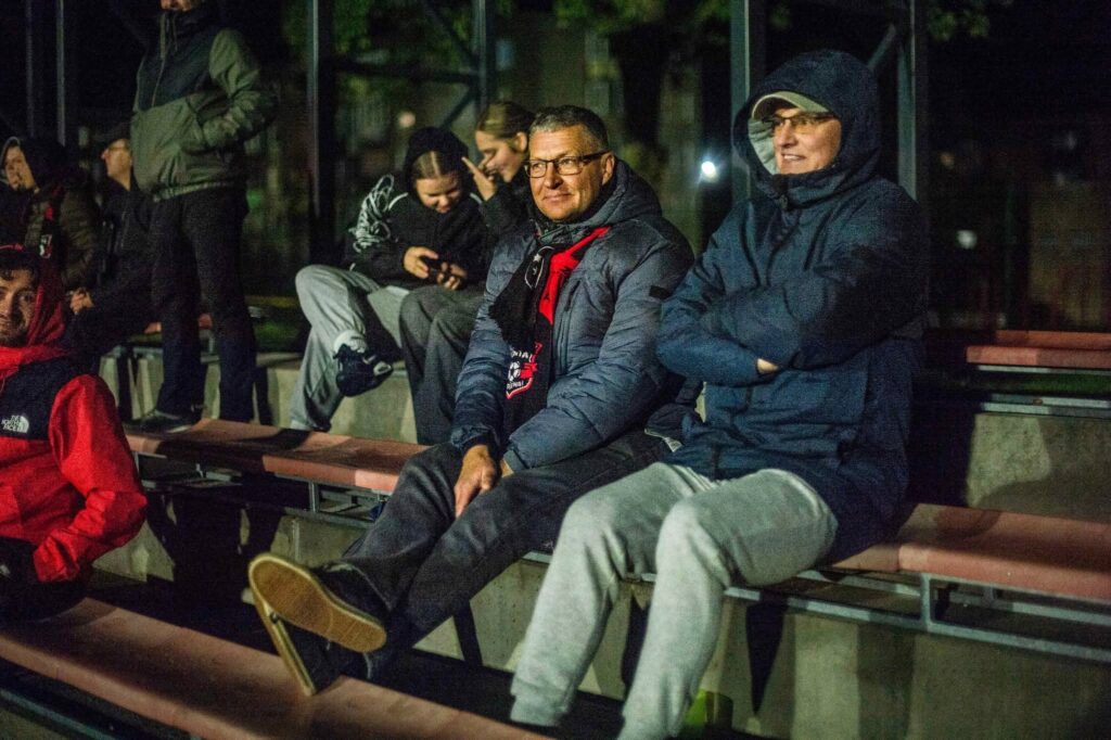 FK „Venta“ rungtynes stebintys žiūrovai vakare sėdi stadiono tribūnose, apsirengę šiltais drabužiais. Pirmame plane – du vyrai su striukėmis, už jų – kiti futbolo sirgaliai.
