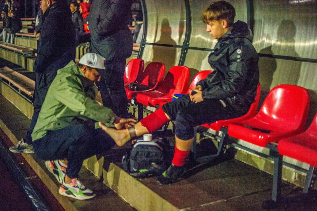 Futbolininkas sėdi ant atsarginių žaidėjų suolelio, o šalia pritūpęs komandos personalo narys apžiūri ir tvarsto jo čiurną rungtynių metu.