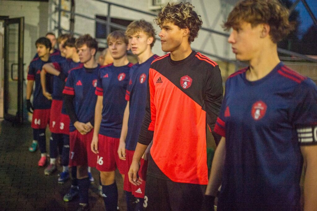 Futbolininkai prieš rungtynes Kuršėnuose. Pirmame plane – ryškiais raudonais ir juodais marškinėliais vilkintis vartininkas, už jo – tamsiai mėlyna ir raudona apranga vilkintys žaidėjai.